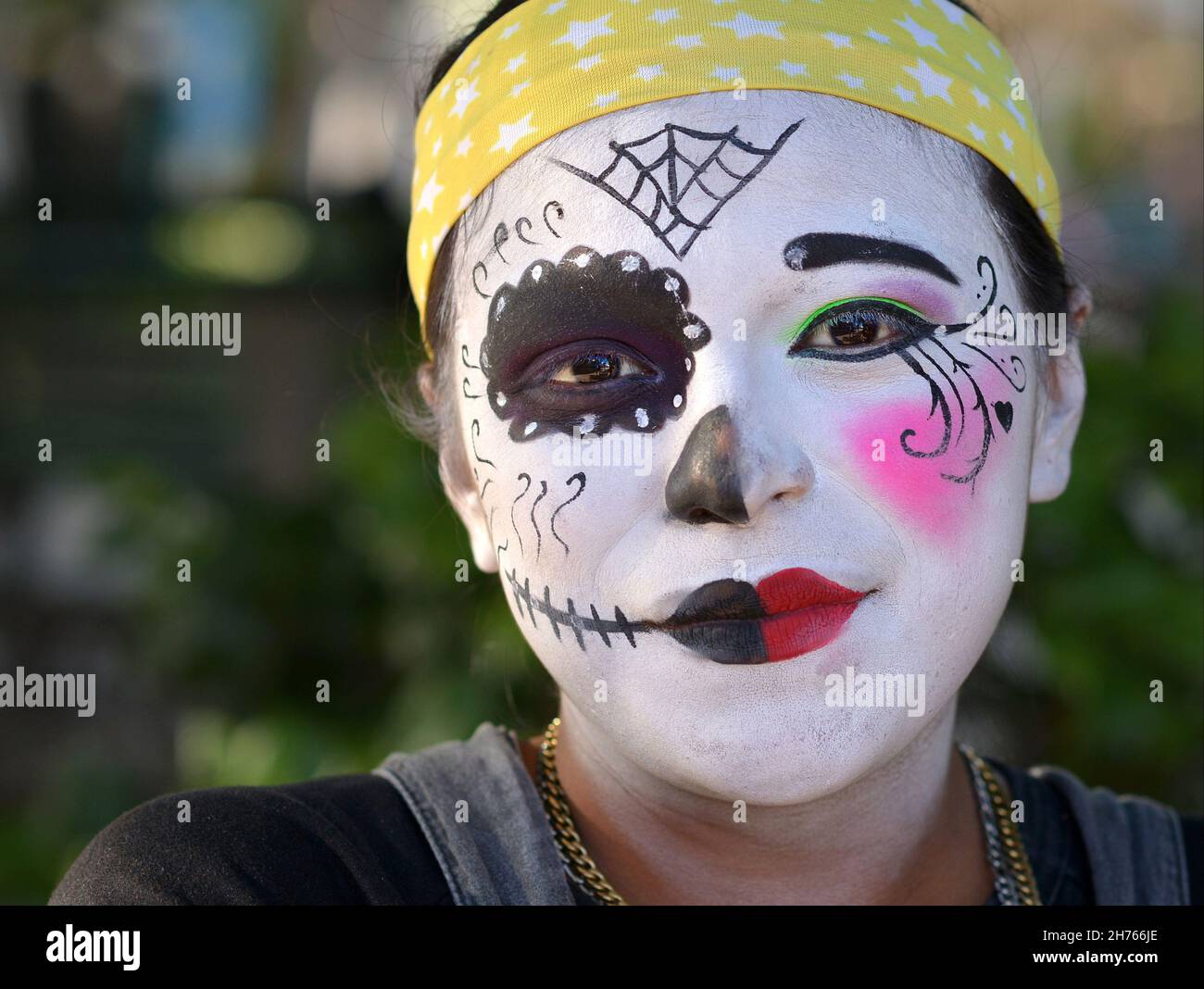 Die junge mexikanische Künstlerin mit zwei catrina- und Clown-Gesichtsmalereien (Tod und Leben) am Tag der Toten (Día de los Muertos) schaut auf den Betrachter. Stockfoto