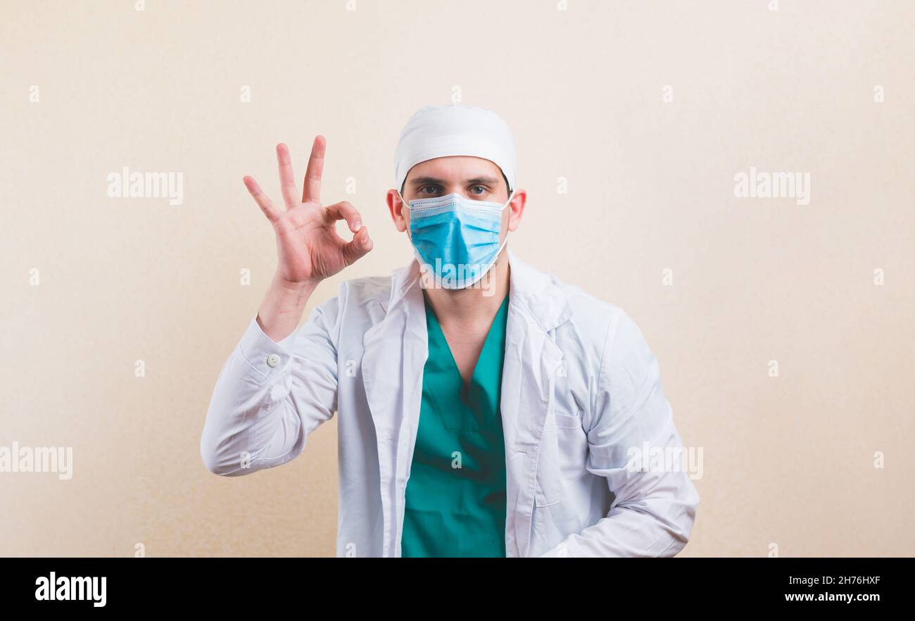 Anwesender Arzt in weißem Anzug und schützender Atemmaske zeigt Friedenszeichen OK bei seinen Händen Stockfoto