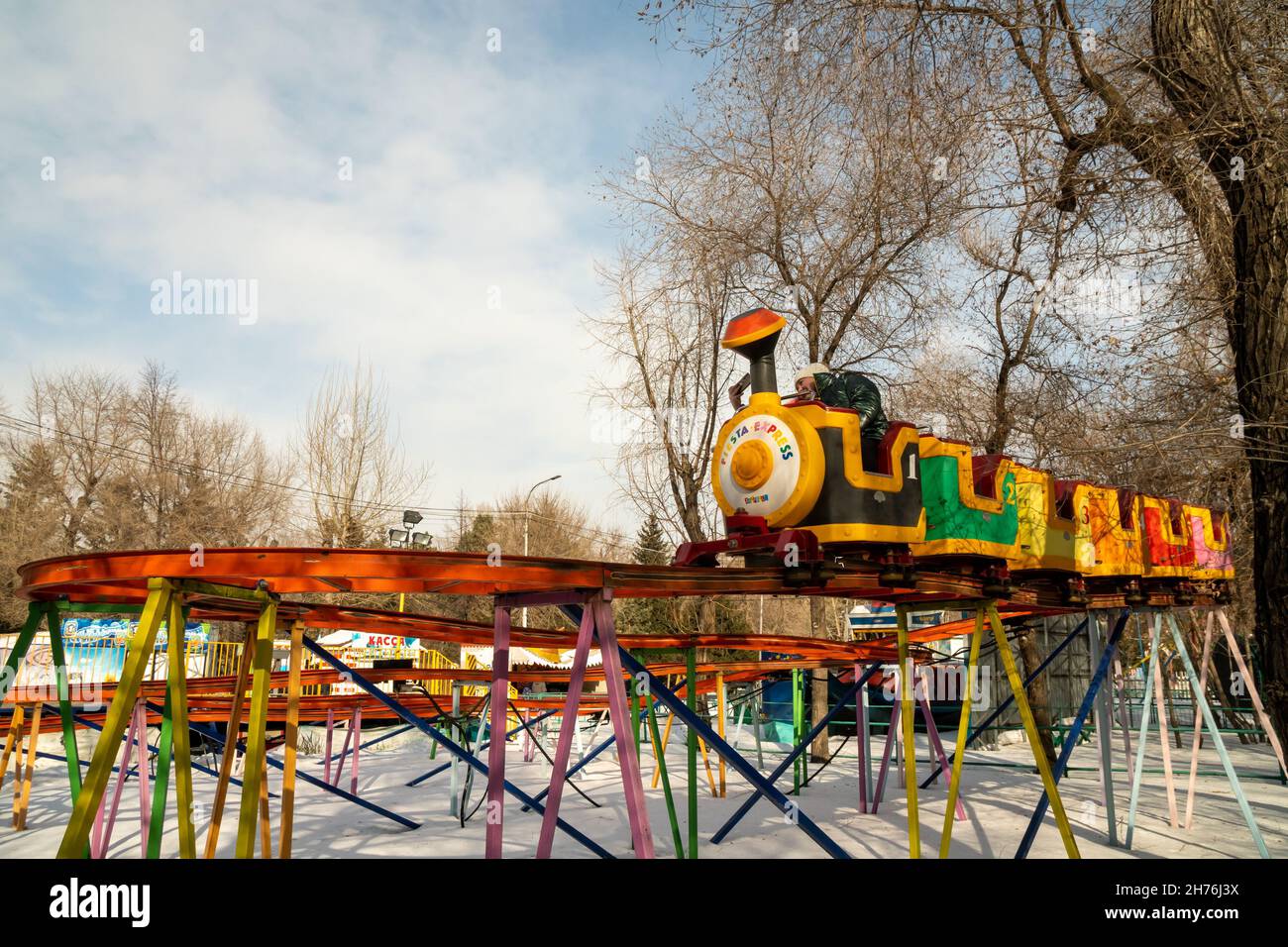 Ein Kinderzug fährt auf hohen Schienen über Schneewehen zwischen Bäumen in einem städtischen Vergnügungspark. Stockfoto