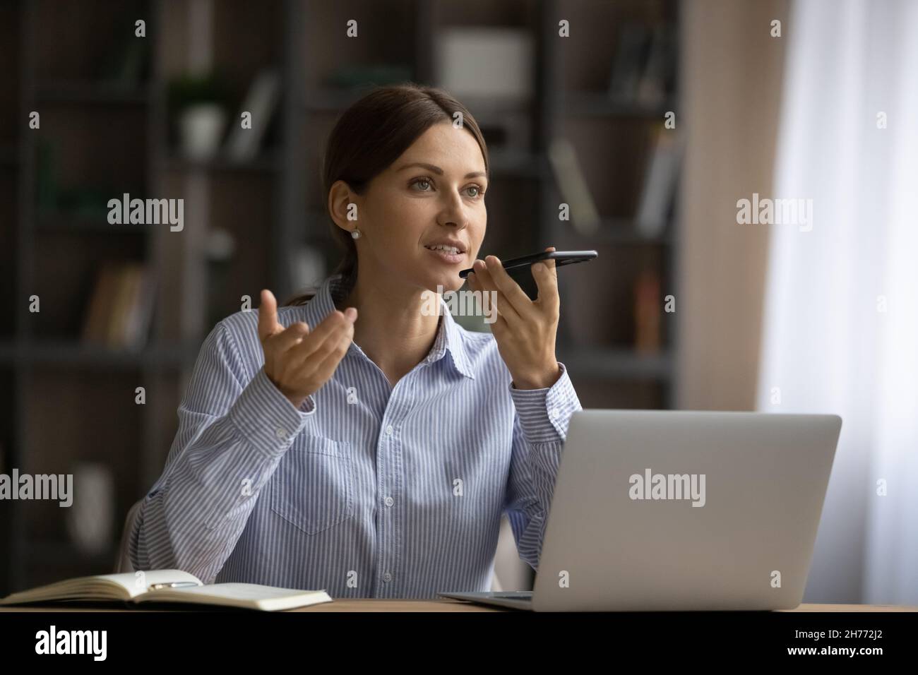 Eine attraktive Frau sitzt am Schreibtisch und hält das Smartphone auf der Freisprecheinrichtung Stockfoto