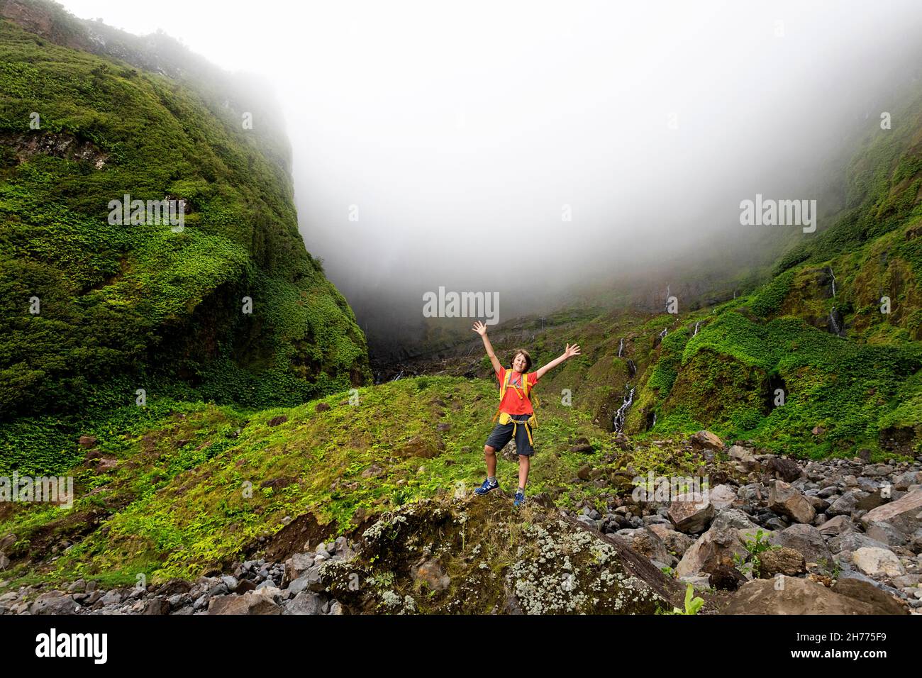 Junge mit einem Rucksack, der lächelt und die Hände beim Ribeira grande Wasserfall hebt. Flores, Azoren, Portugal Stockfoto