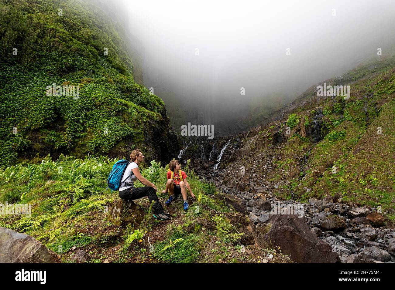 Mutter und Sohn mit Rucksäcken sitzen auf Felsen am Fuße des Ribeira grande Wasserfalls im Nebel. Flores, Azoren, Portugal Stockfoto