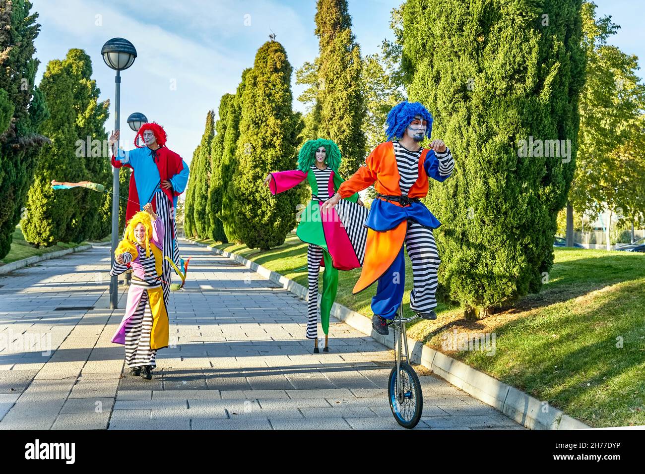 Ganzkörper-Truppe mit lustigen, kreativen Künstlern in bunten Harlekinen-Kostümen und Perücken, die an sonnigen Tagen im Park auf Stelzen spazieren und mit einem Einrad fahren Stockfoto