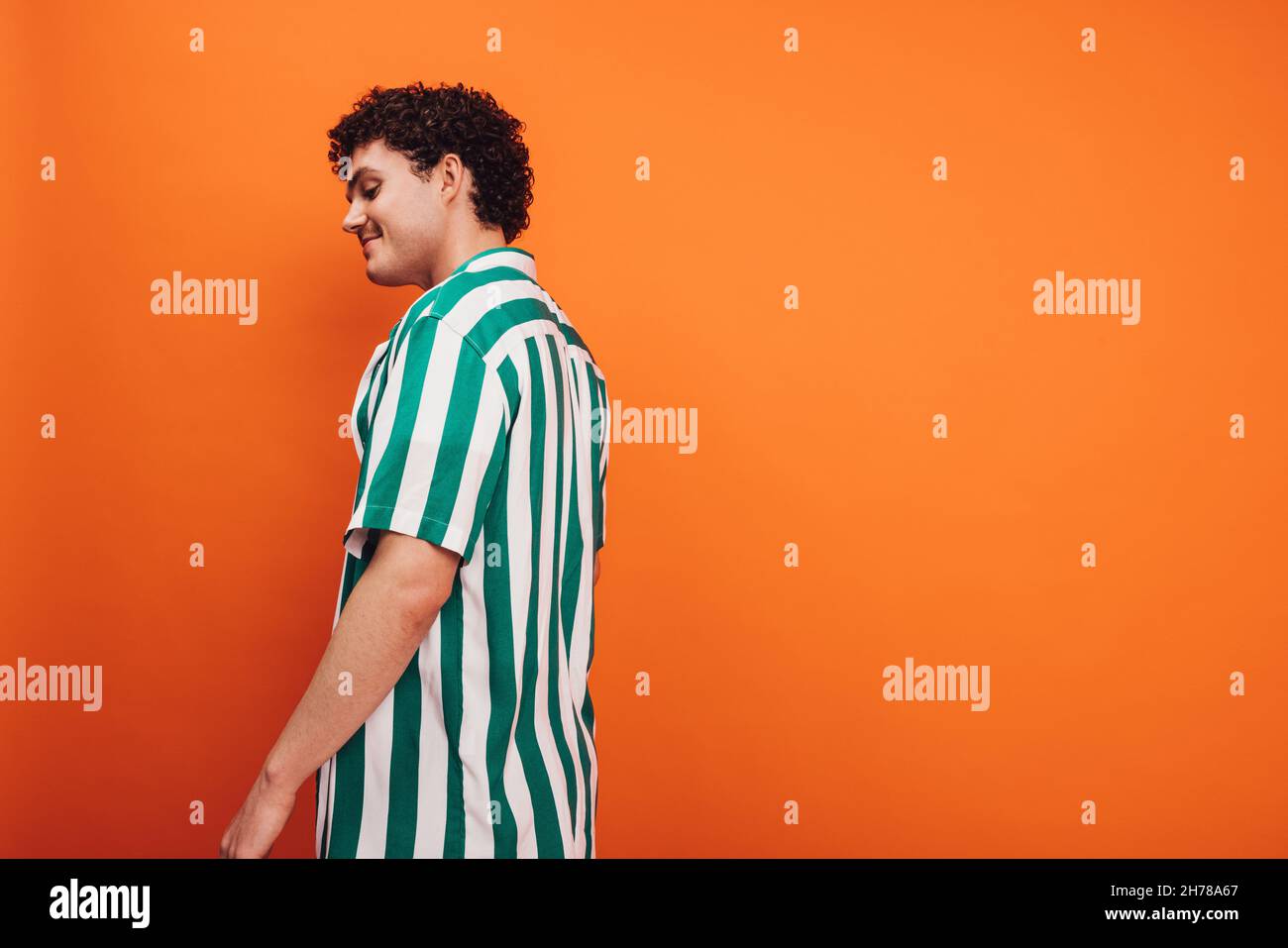 Fröhlicher junger queerer Mann, der vor einem orangefarbenen Hintergrund steht. Seitenansicht eines glücklichen jungen Generation z Hipsters, der selbstbewusst im Stehen aussieht Stockfoto