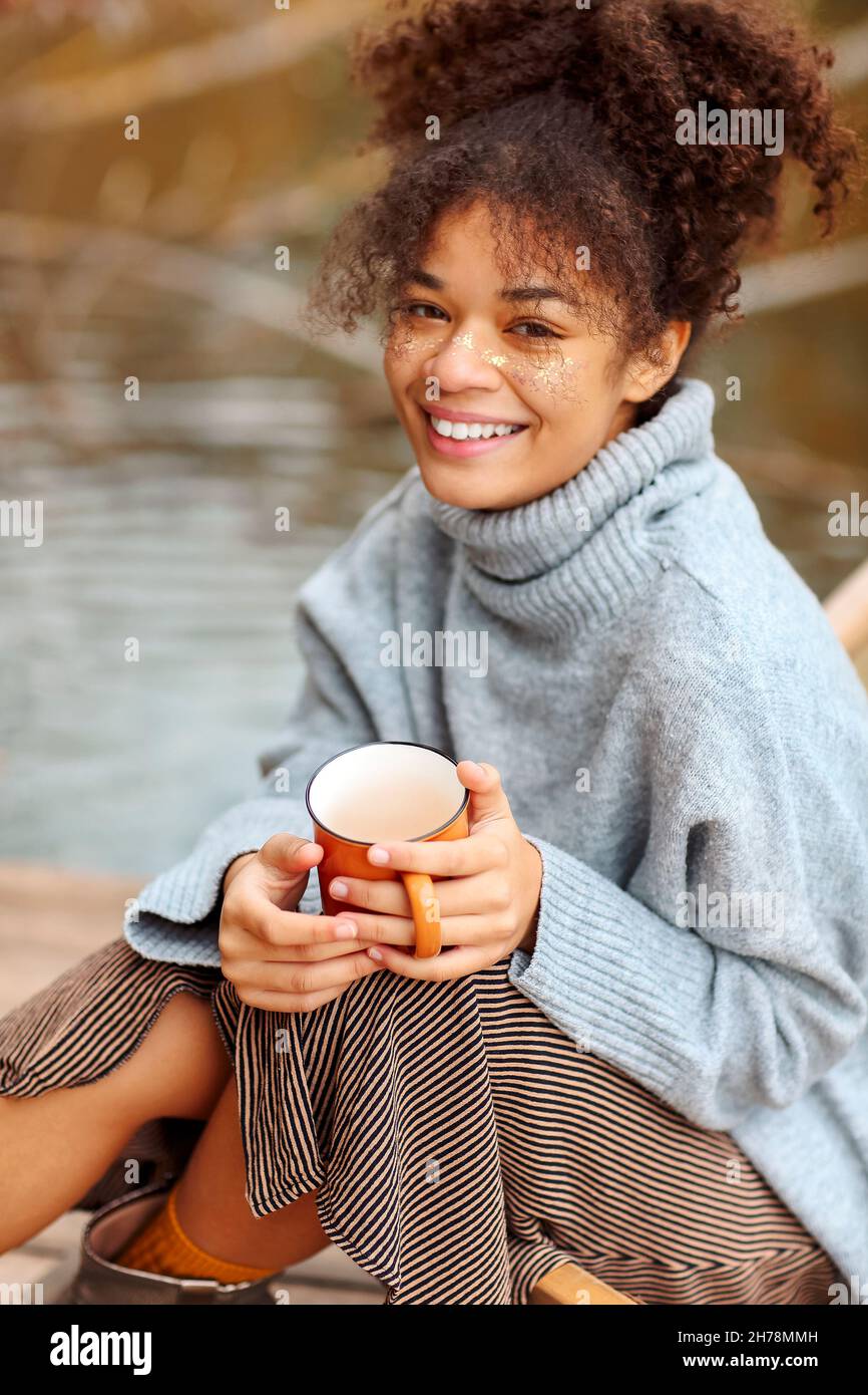 Junge glückliche, sorglose afroamerikanische Frau mit einer Tasse heißen Tee, die sich im Herbst in der Nähe des Teiches entspannt, Mischrasse-Weibchen in warmem Pullover, die im Herbst die Wehe genießen Stockfoto