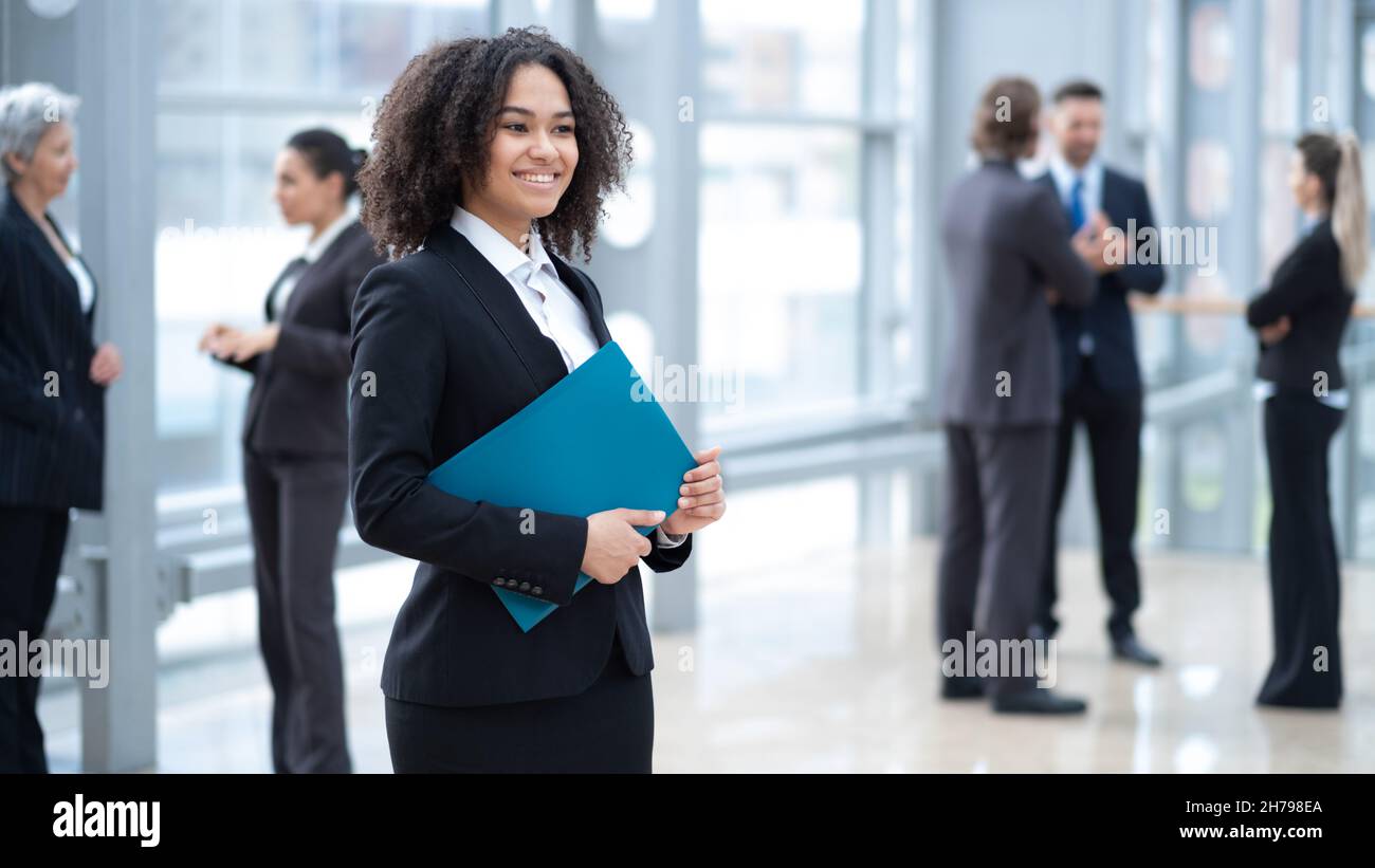 Nahaufnahme eines Porträts einer jungen Geschäftsfrau mit gemischter Rasse, die mit einem Dokumentenordner vor Kollegen im Geschäftsgebäude steht Stockfoto