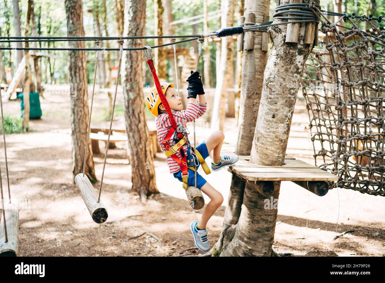 Boy in einem Sicherheitsgurt klettert auf eine Baumplattform über die Agility Bridge Stockfoto