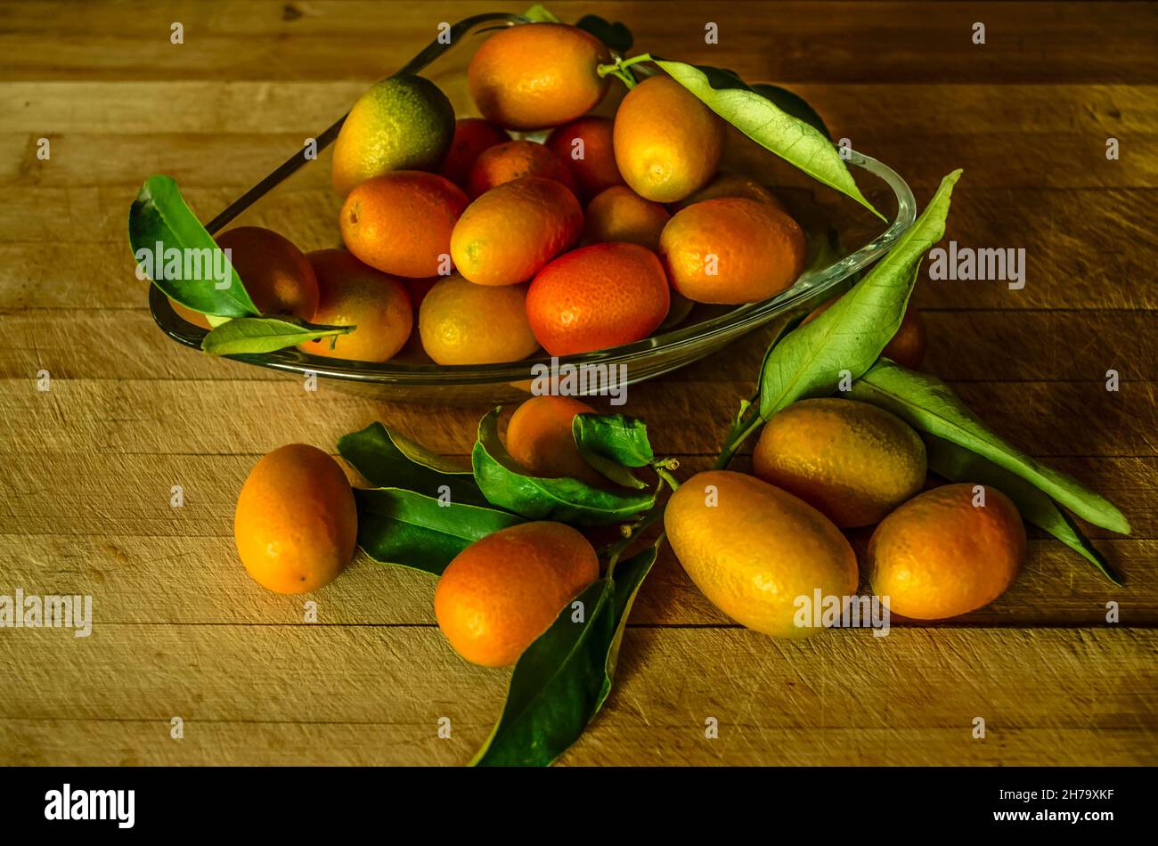 Kleine Glasplatte mit kleinen Kumquat-Orangen mit grün glänzenden Blättern und einer Gruppe von Kumquat-Orangen, die in der Nähe auf einem Holztisch liegen Stockfoto