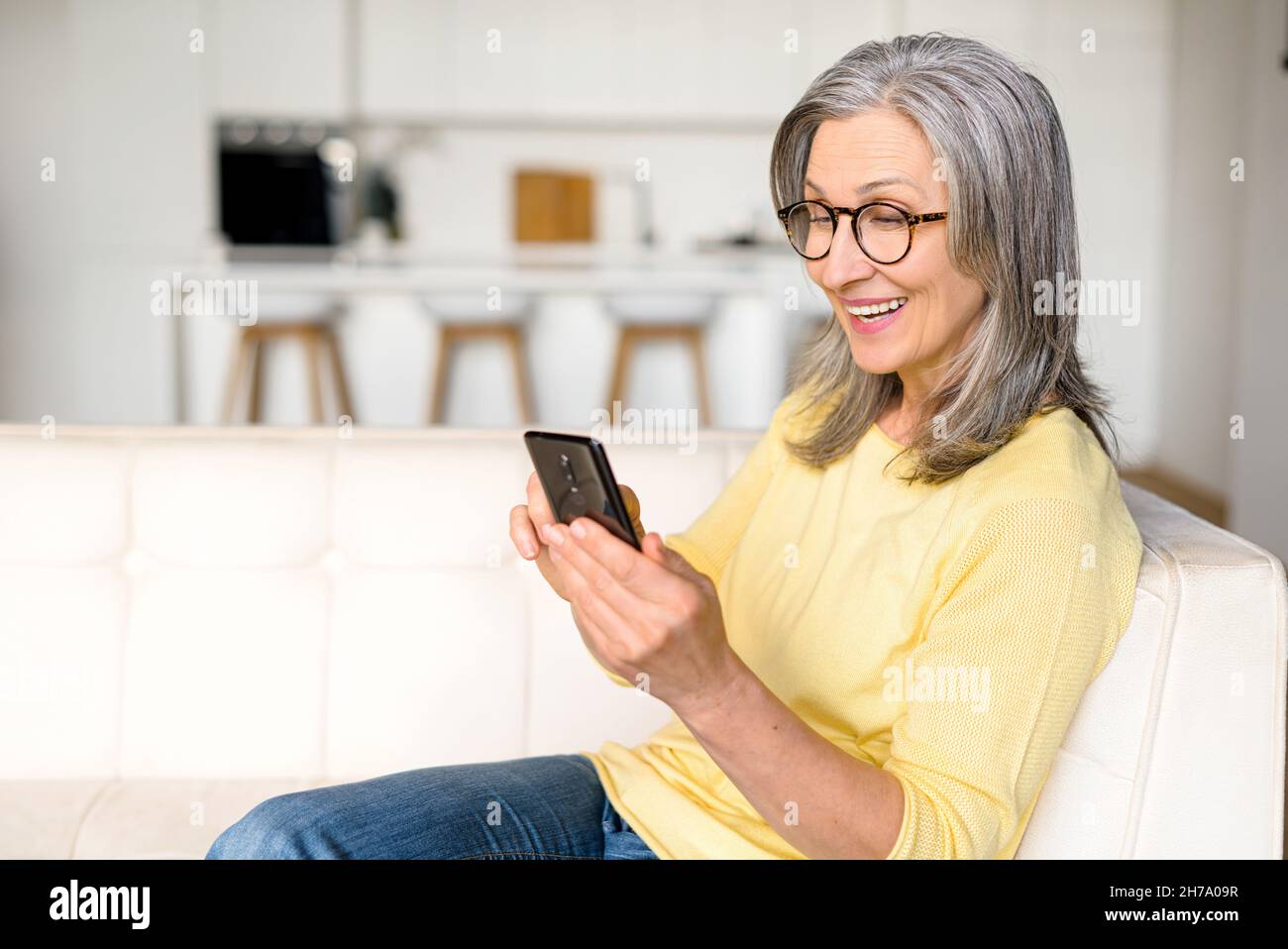 Positive ältere Dame, die in entspannter Pose auf der bequemen Couch mit einem Smartphone sitzt, verbringt eine ältere grauhaarige Frau ihre Freizeit damit, in den Neuigkeiten zu blättern und im Internet zu surfen Stockfoto