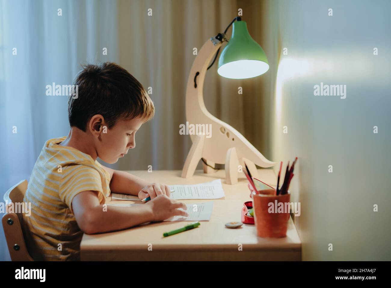 Junge macht seine Hausaufgaben, während er am Schreibtisch sitzt. Home interier.DIY Dinosaurier-Lampe beleuchtet die Tischplatte Stockfoto