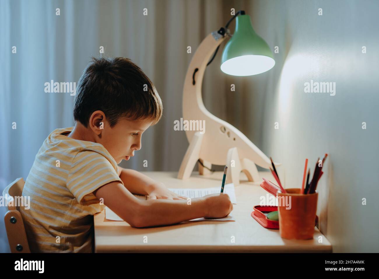 Junge macht seine Hausaufgaben, während er am Schreibtisch sitzt. Home interier.DIY Dinosaurier-Lampe beleuchtet die Tischplatte Stockfoto