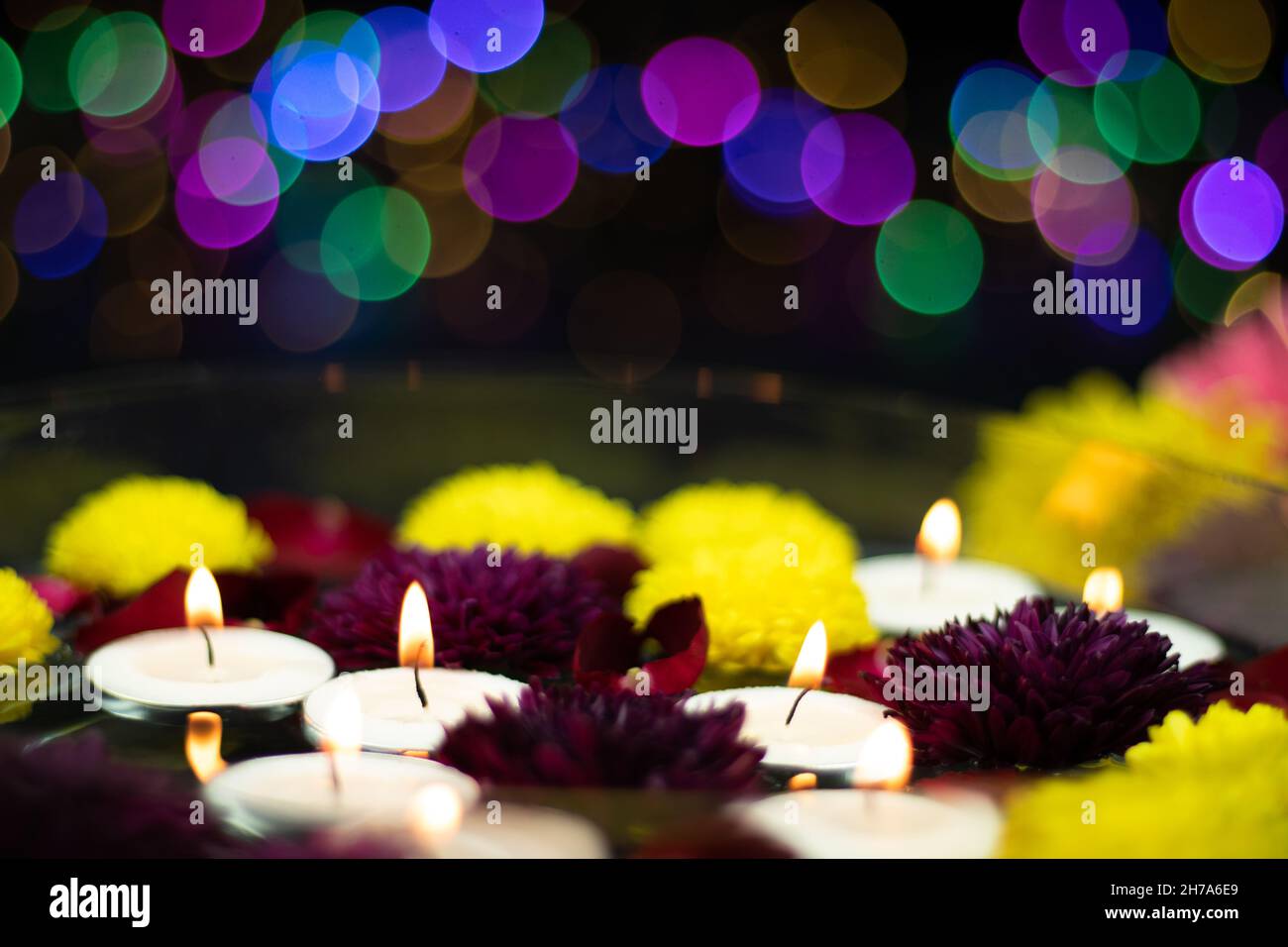 Schwimmende Teelichtkerzen, Die Von Frischen Bunten Maroon- Und Gelben Blumen Umgeben Sind. Buntes Bokeh Auf Dunklem Hintergrund. Thema Für Shubh Stockfoto