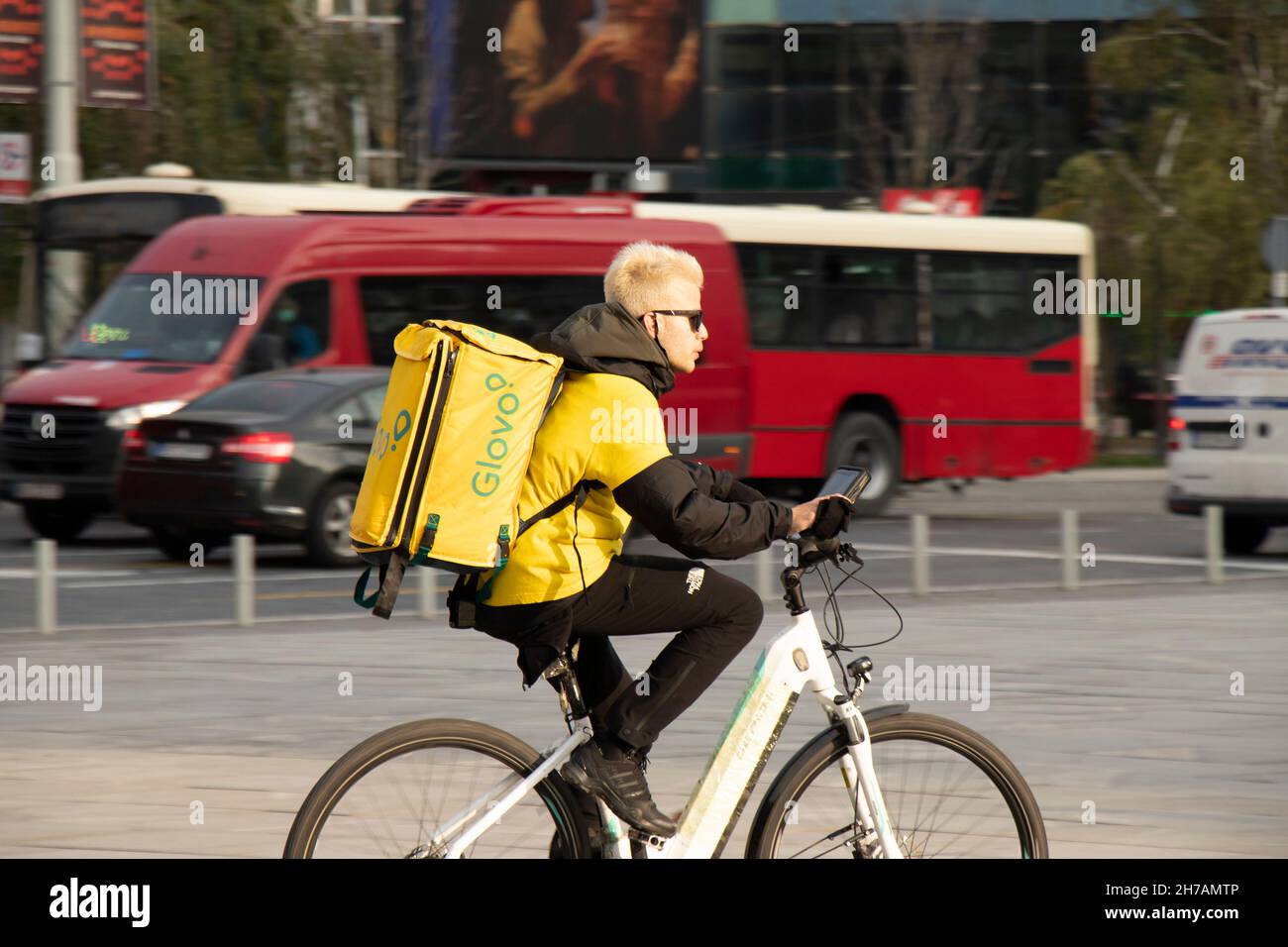 Belgrad, Serbien - 17. November 2021: Junger Mann, der für den Lebensmittellieferdienst der Stadt Glovo arbeitet, fährt mit dem Fahrrad über den Stadtplatz Stockfoto