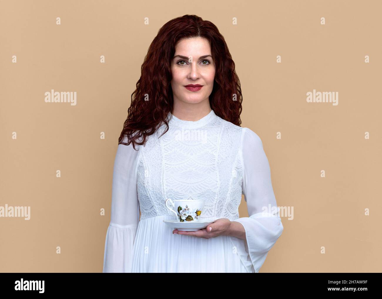 Frau hält Teetacup mit weichem Lächeln auf beigem Hintergrund. Porträt einer schönen Frau in weißem Kleid mit einem Vintage-Teetasse mit Rose-Design. Stockfoto