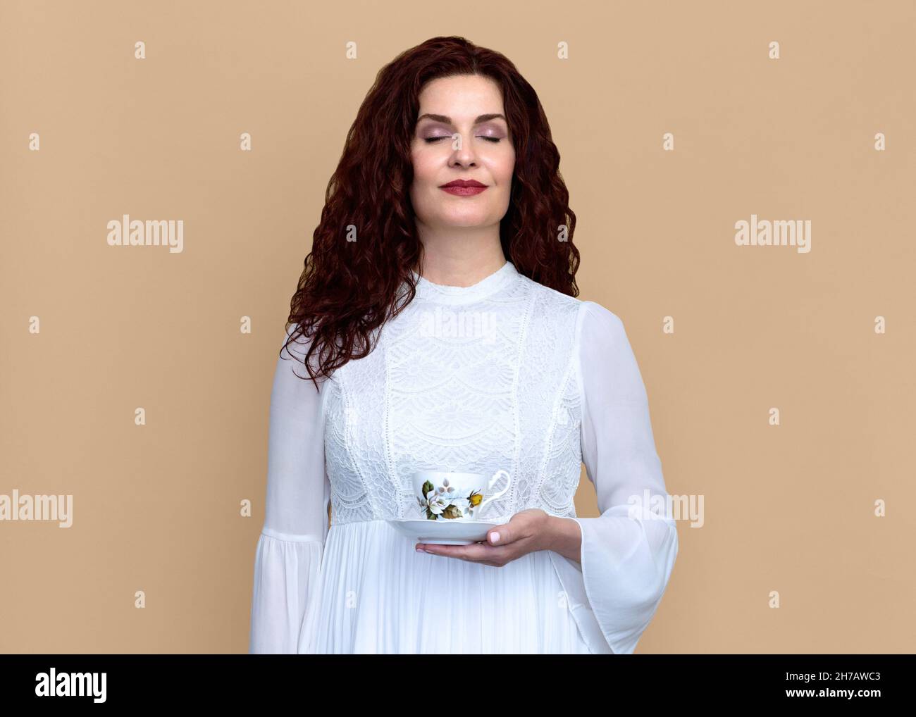 Frau mit geschlossenen Augen hält und riecht Tee mit Beige Hintergrund. Sie trägt ein romantisches weißes Kleid und hat welliges rotbraunes Haar und ein weiches Lächeln. Stockfoto