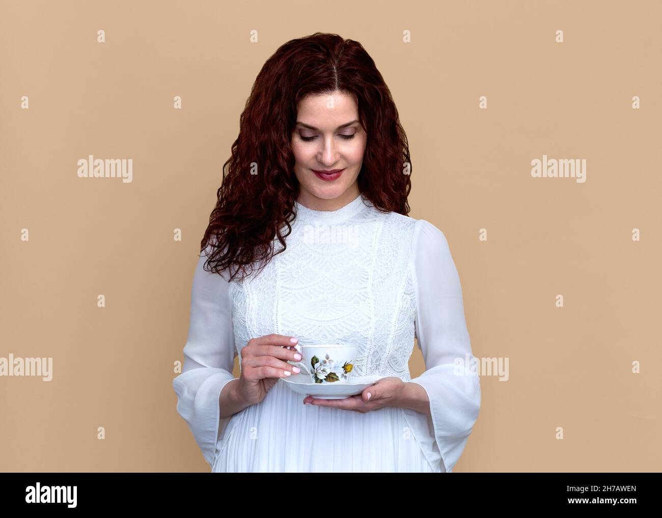 Frau schaut nach unten Halten feine Knochen China Teetacup mit Beige Hintergrund. Schöne lächelnde Frau in romantischen weißen Kleid hält vintage Teetasse. Stockfoto