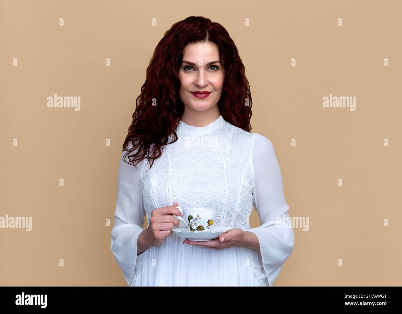 Frau mit weichem Lächeln hält Teetasse isoliert gegen Beige Hintergrund schön lächelnde Frau in romantischen weißen Kleid hält vintage Teetasse. Stockfoto