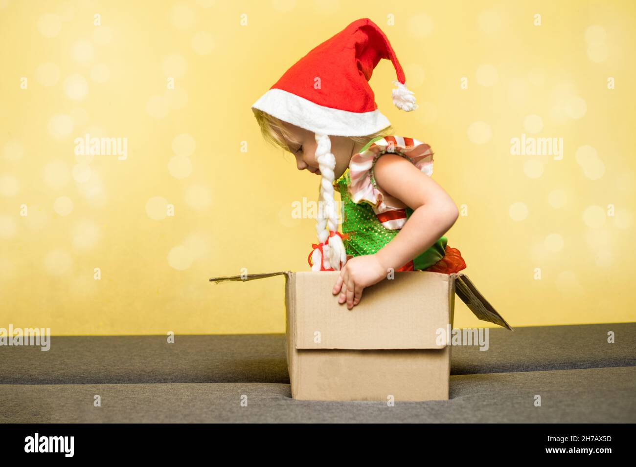 Kleines Mädchen in Santa's Hut und ein elfenbegleites Kostüm sitzt in der Karton-Box. Konzept der Empfang oder Versand Weihnachtspäckchen zu Hause. Lieferung ser Stockfoto