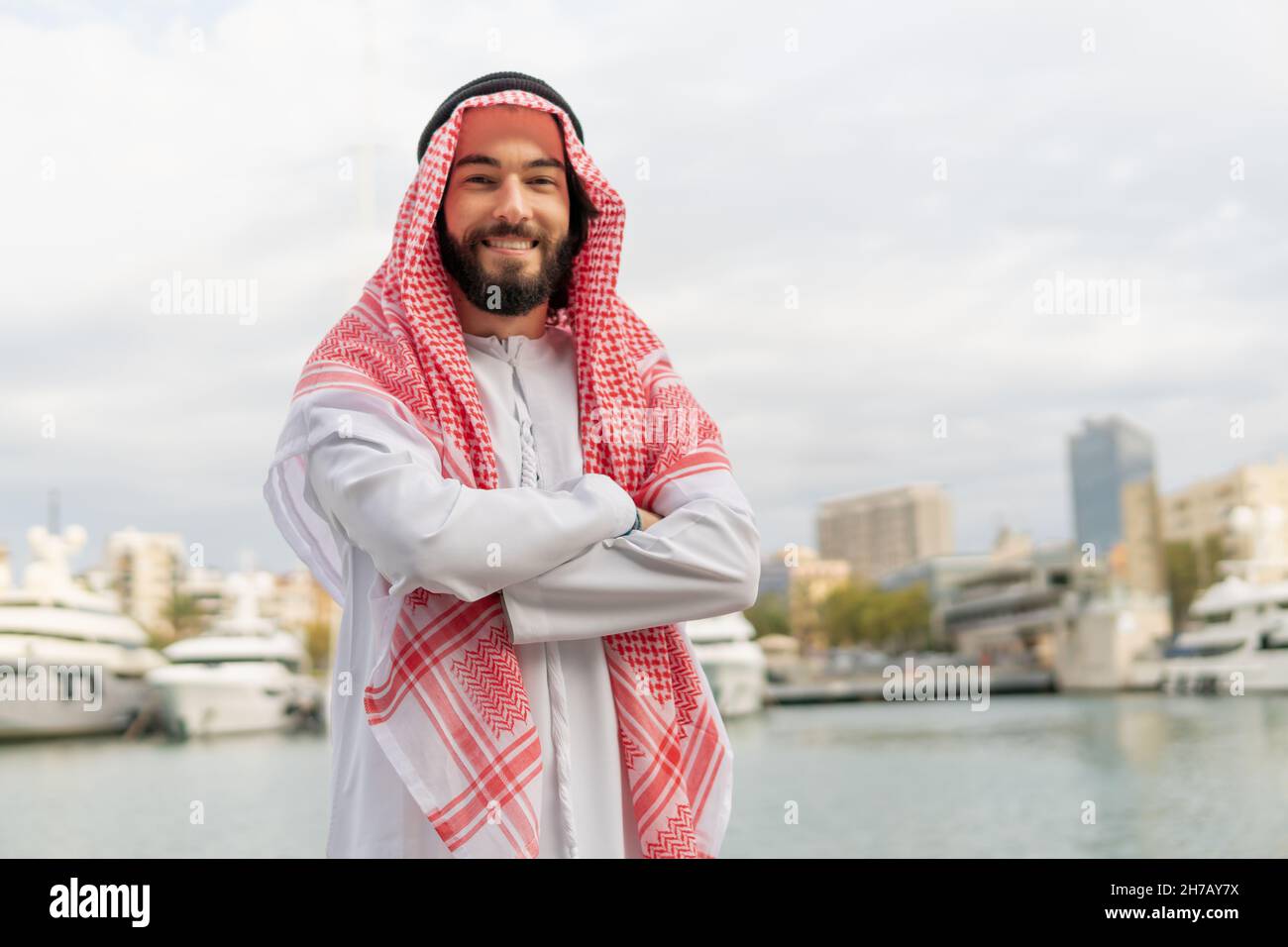 Porträt eines fröhlichen, fröhlichen arabischen Mannes in traditioneller Kleidung, keffiyeh und weißer Thobe, der die Kamera anlächelt, während er im Hafen steht und die Arme gekreuzt hält. Foto des erfolgreichen saudischen Geschäftsmannes Stockfoto