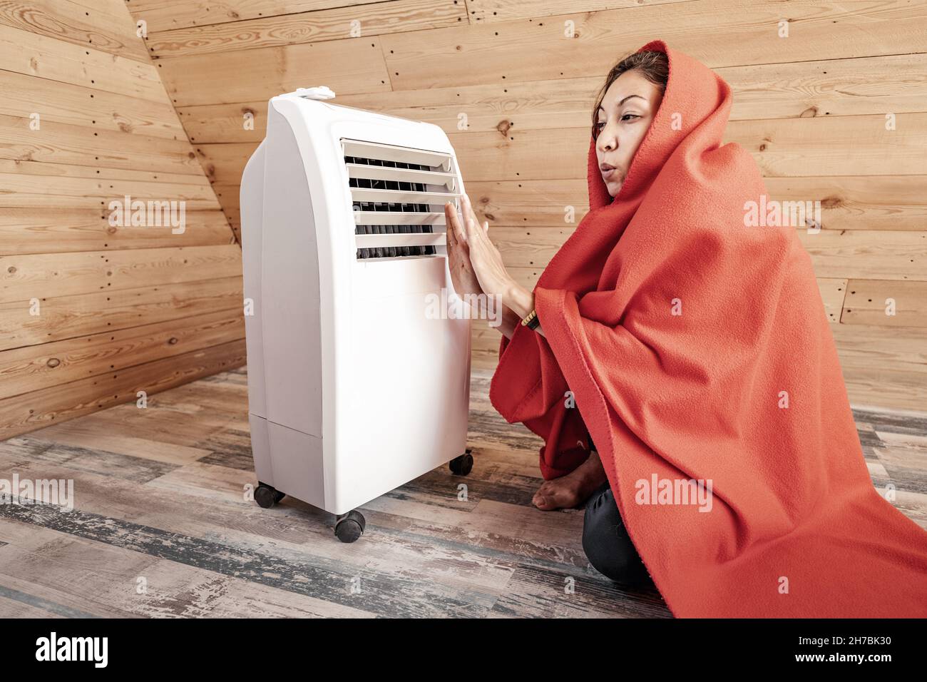 Eine lustige Frau wärmt sich im Winter an einem elektrischen Heizkörper in ihrem Holzhaus auf, eingewickelt in eine Decke. Das Konzept von Energie und Komfort in der kalten Jahreszeit Stockfoto