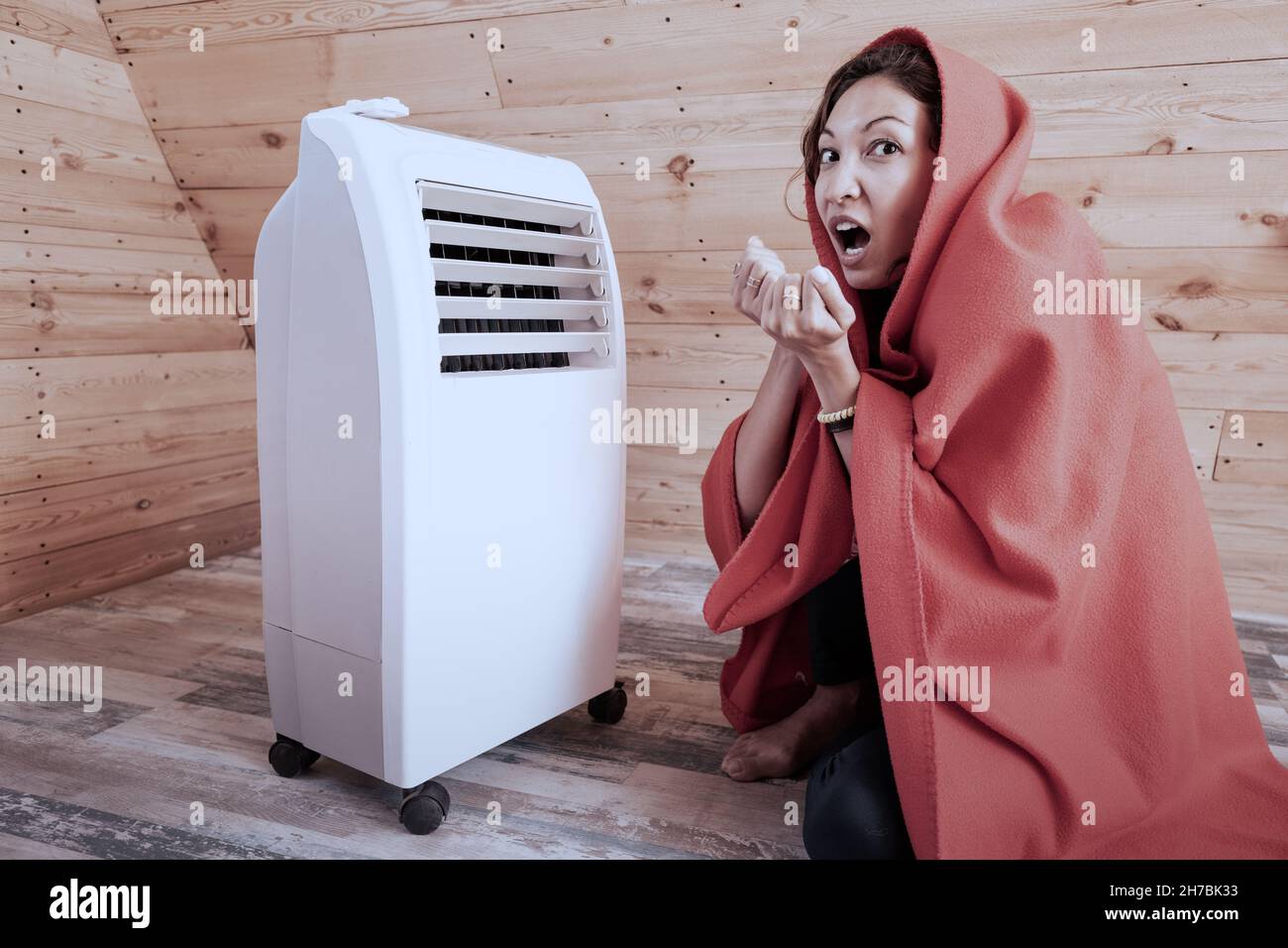 Lustige Frau, die im Winter in ihrem Holzhaus in der Nähe eines elektrischen Heizkörperes friert, eingewickelt in eine Decke. Das Konzept von Energie und Komfort in der kalten Jahreszeit. Stockfoto