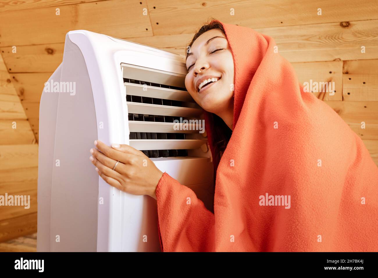 Eine lustige Frau wärmt sich im Winter an einem elektrischen Heizkörper in ihrem Holzhaus auf, eingewickelt in eine Decke. Das Konzept von Energie und Komfort in der kalten Jahreszeit Stockfoto