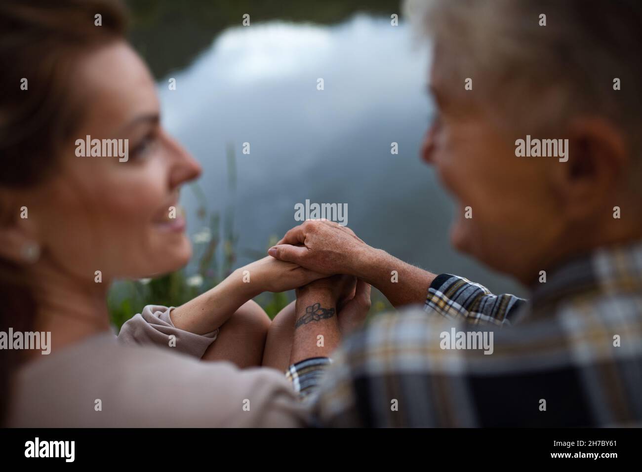 Rückansicht der älteren Mutter, die die Hände hält, während die Erwachsene Tochter in der Natur am See sitzt, und auf die Hände konzentrieren Stockfoto