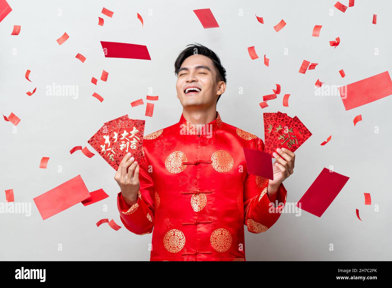 Glücklich schöner asiatischer Mann in traditioneller orientalischer Tracht mit roten Umschlägen oder Ang Pao auf grauem Hintergrund mit Konfetti, chinesische Texte bedeuten großartig Stockfoto