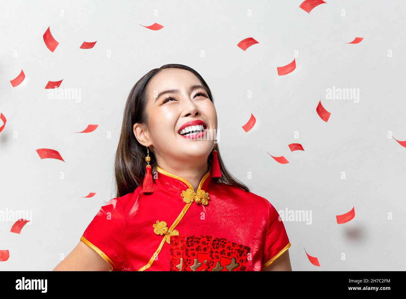 Lächelnde glückliche asiatische Frau in traditioneller orientalischer Tracht, die auf hellgrauem Hintergrund mit rotem Konfetti nach oben blickt Stockfoto