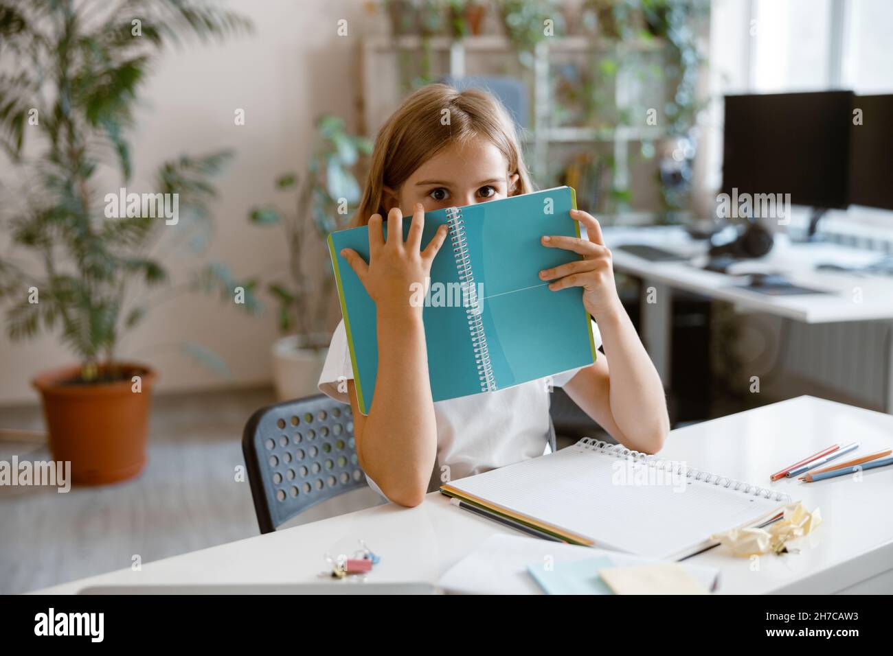 Schockiertes kleines Mädchen versteckt sich hinter Notebook am Tisch im hellen Raum Stockfoto