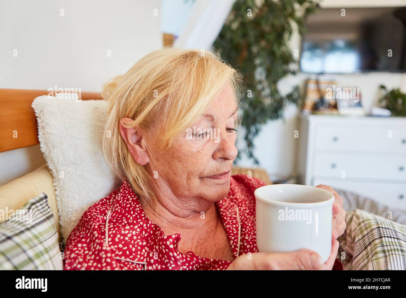 Die alte Frau liegt traurig und nachdenklich im Bett zu Hause und trinkt eine Tasse Kaffee Stockfoto