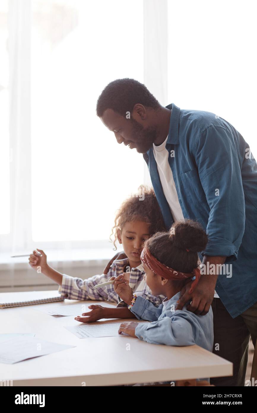 Vertikale Seitenansicht Porträt eines fürsorglichen afroamerikanischen Vaters, der Hausaufgaben mit zwei kleinen Mädchen zu Hause macht Stockfoto