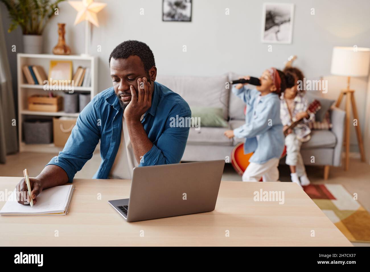 Porträt eines afroamerikanischen Mannes, der einen Laptop benutzt, während er zu Hause mit Kindern arbeitet, die im Hintergrund spielen, und Kopierraum Stockfoto