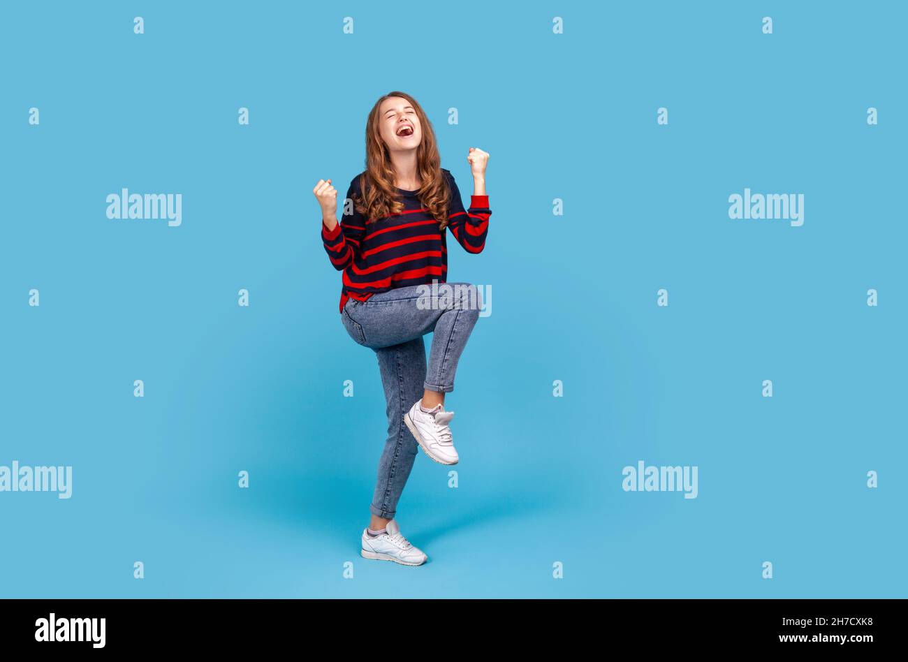 Durchgehende positive zufriedene Frau trägt gestreiften lässigen Pullover und Jeans, erhöhte geballte Fäuste, glücklich über den Sieg, zum Ausdruck bringen Triumph. Innenaufnahme des Studios isoliert auf blauem Hintergrund. Stockfoto