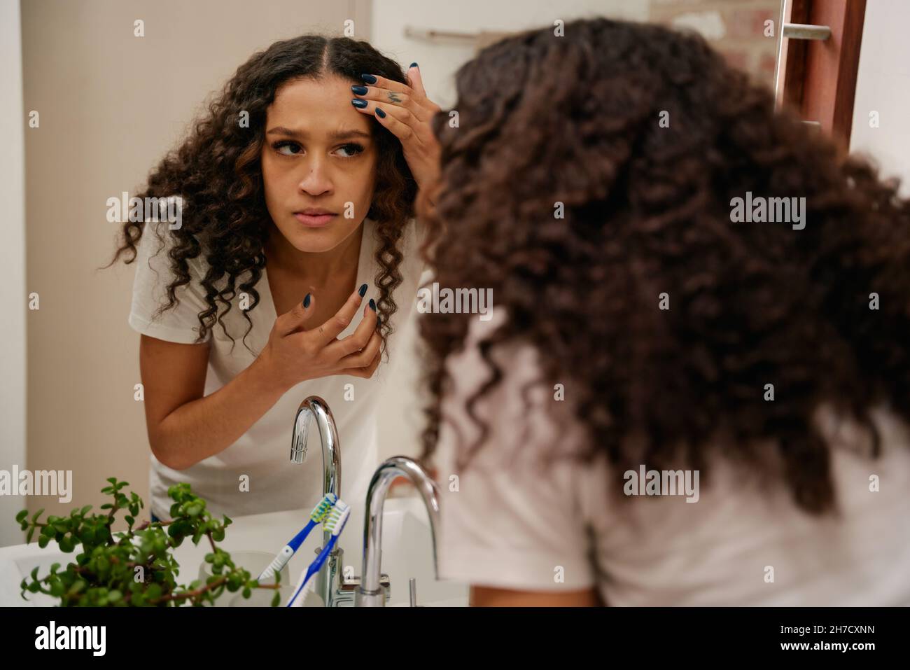 Multikulturelle junge Frau, die besorgt auf ihre Haut im Spiegel im modernen Badezimmer schaut Stockfoto