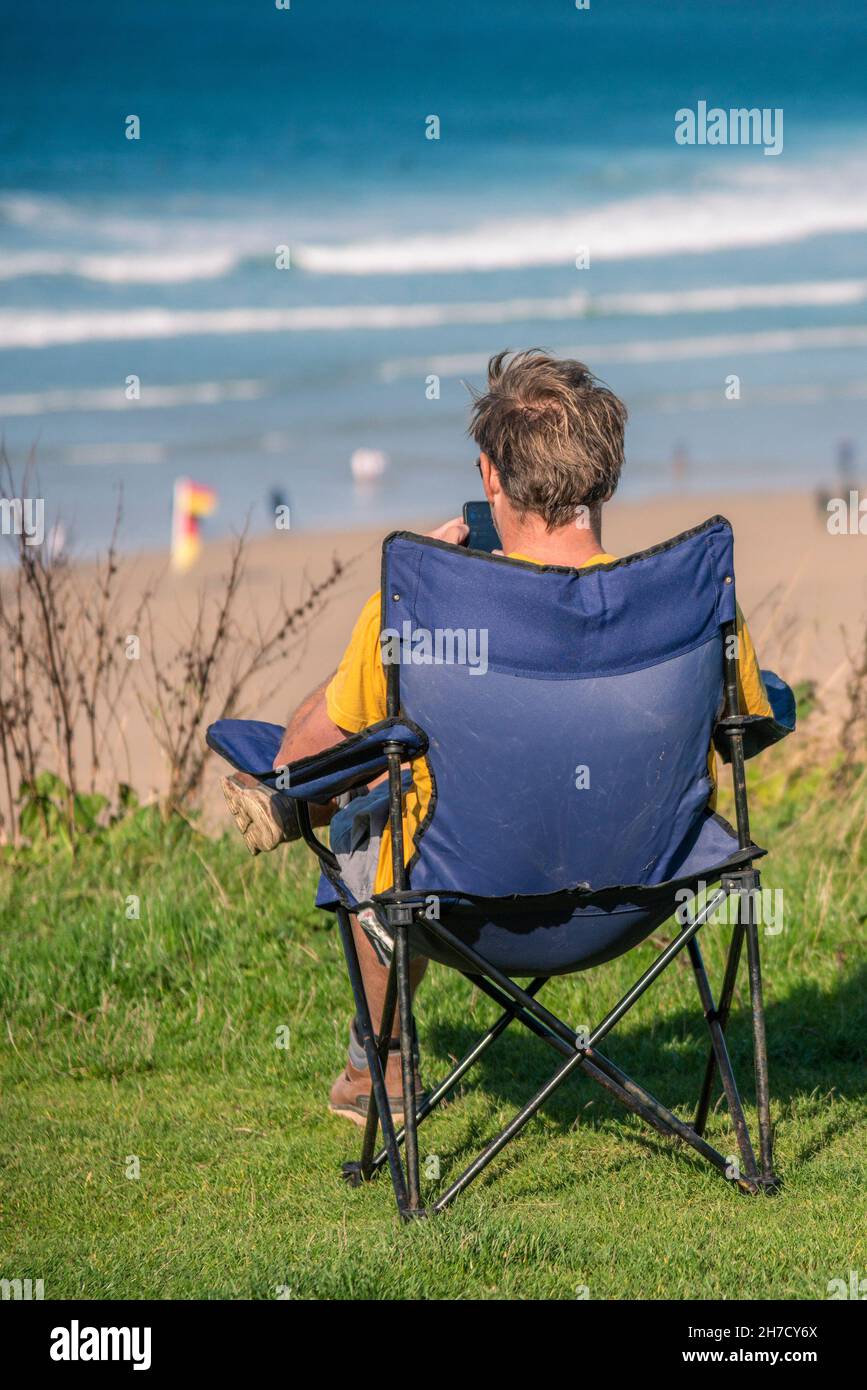 Ein männlicher Urlauber sitzt und entspannt in einem Campingstuhl mit Blick auf Fistral Beach in Newquay in Cornwall. Stockfoto