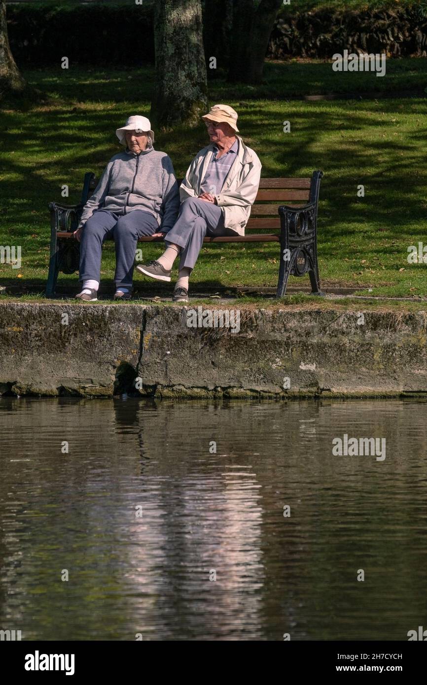 Ein älteres Paar, das einen Plausch und die warme Sonne genießt, die auf einer Bank in der Nähe eines Sees in einem Park sitzt. Stockfoto