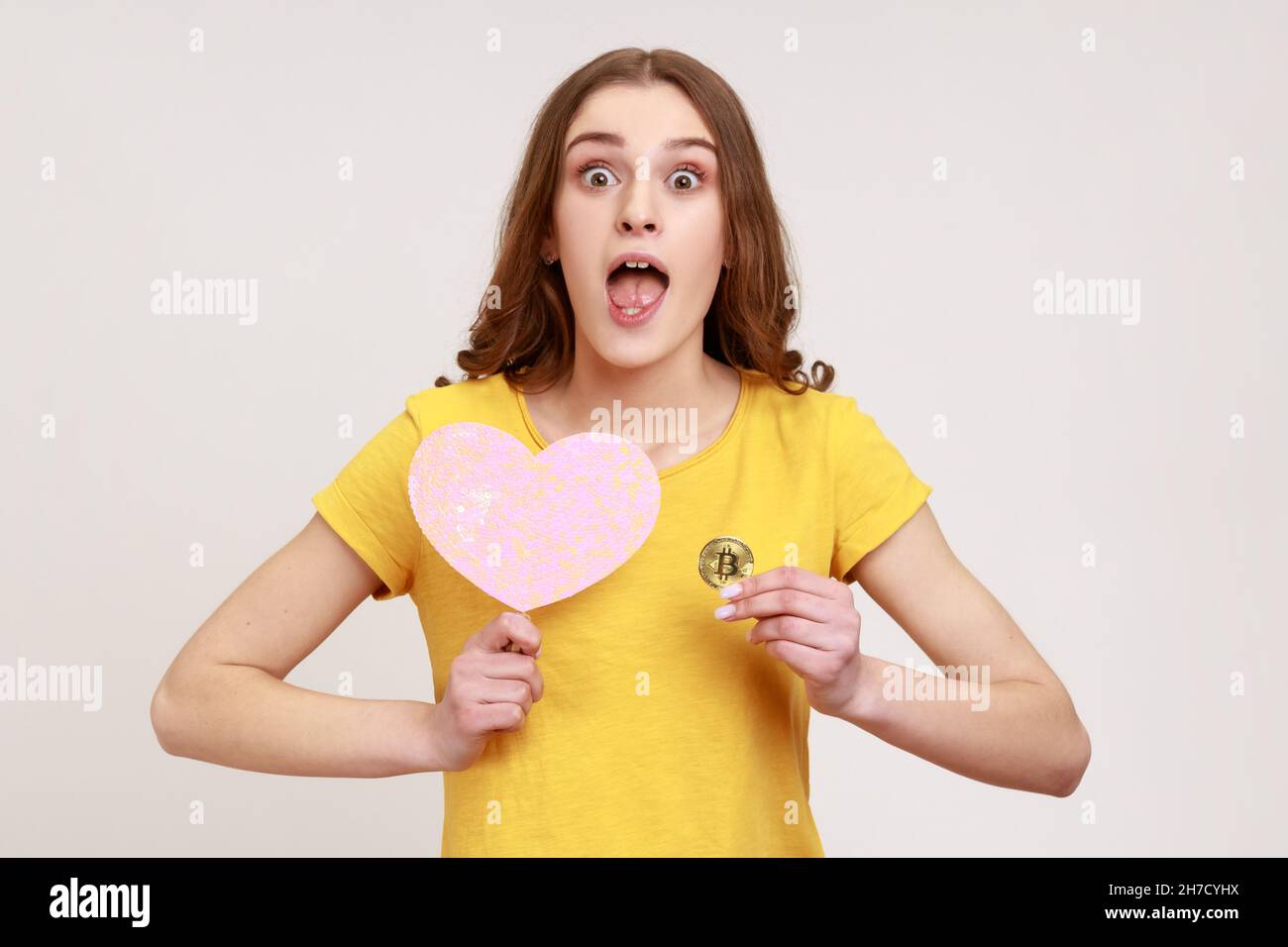 Porträt der jungen überraschten Frau in gelben casual T-Shirt mit rosa Herz auf Stick und physischen Bitcoin, hat Gesichtsausdruck schockiert, Kryptowährung. Innenaufnahme des Studios isoliert auf grauem Hintergrund Stockfoto