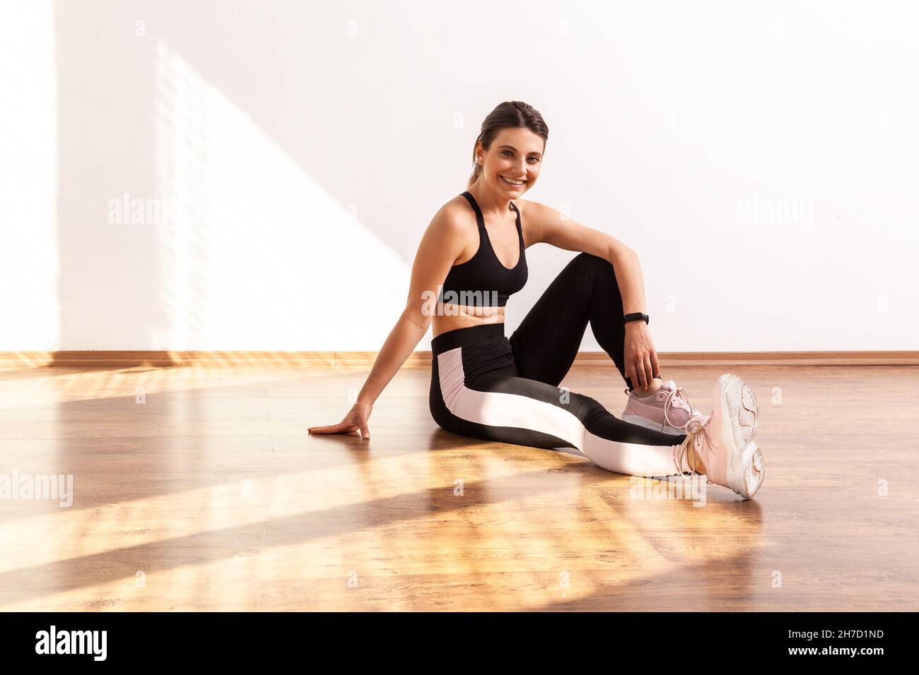 Frau, die auf dem Boden sitzt, nach dem Training entspannt Pause macht, sich nach dem Training attraktiv fit fühlt, schwarzes Sporttop und Strumpfhosen trägt. Studioaufnahme in voller Länge, die durch Sonnenlicht vom Fenster beleuchtet wird. Stockfoto