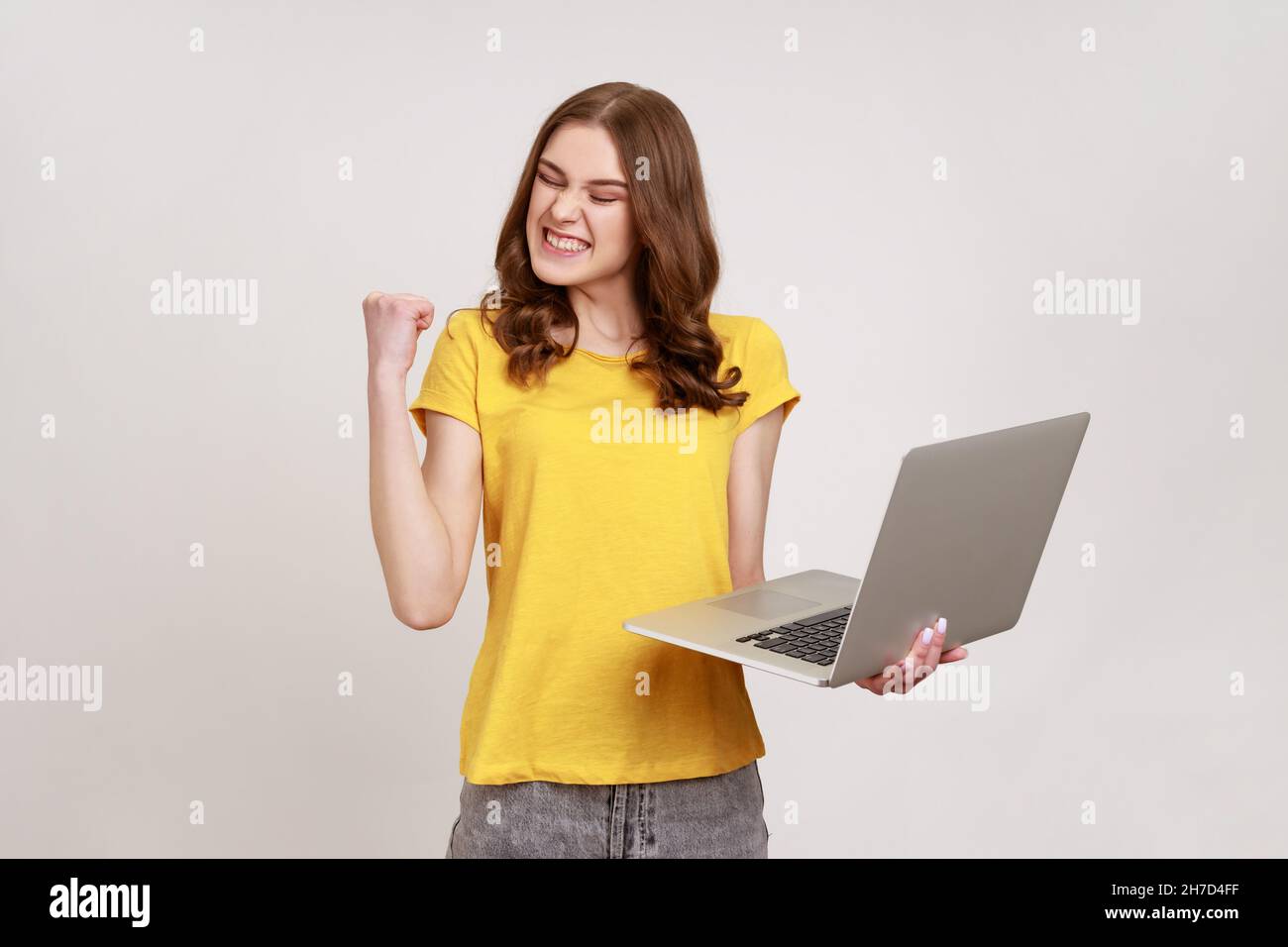 Portrait von begeisterten überglücklich Teenager Mädchen in gelben T-Shirt mit Faust und Laptop halten, jubeln Sieg, Online-Wetten. Innenaufnahme des Studios isoliert auf grauem Hintergrund. Stockfoto