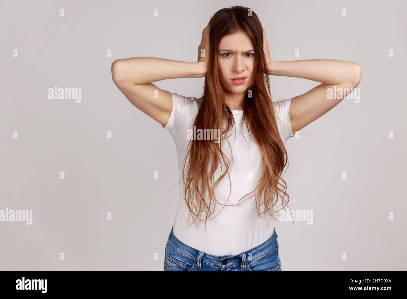 Gereizte gestresste Frau, die Ohren bedeckt und mit verzweifeltem, verärgertem Ausdruck aussieht, Stimmen hört, Nervenzusammenbruch, weißes T-Shirt trägt. Innenaufnahme des Studios isoliert auf grauem Hintergrund. Stockfoto