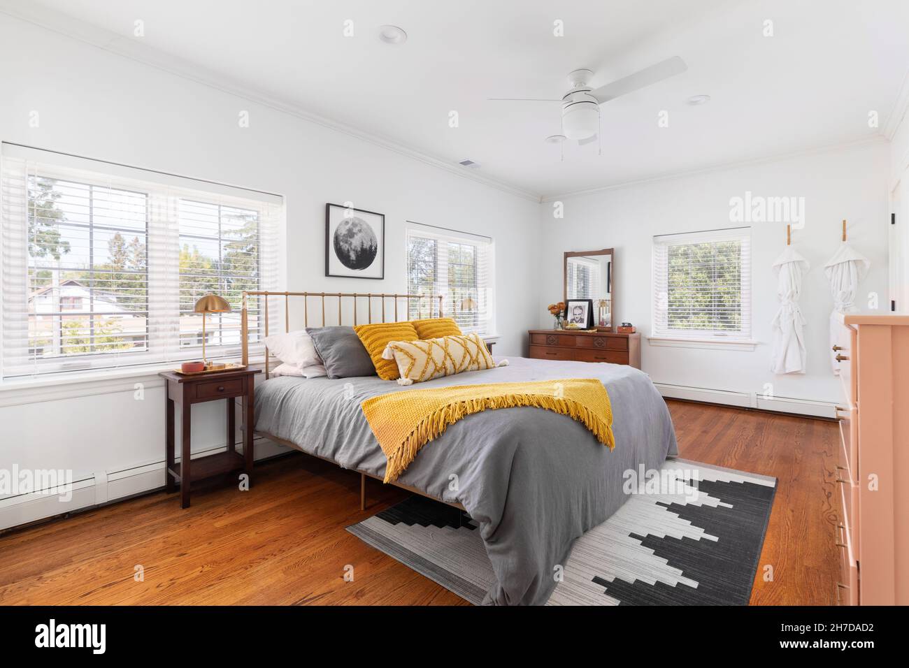 Ein helles Schlafzimmer mit weißen Wänden, gelb-grauer Bettwäsche und einem Teppich auf Hartholzböden. Stockfoto