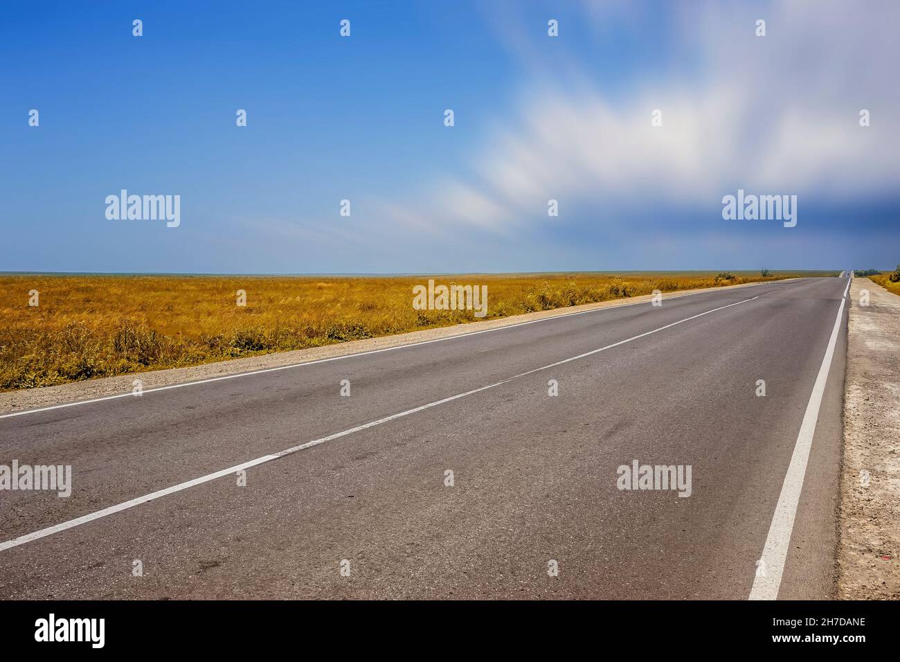 Eine lange Autobahn ohne Autos auf dem überwucherten Gras der Steppe unter einem blau bewölkten Himmel Stockfoto