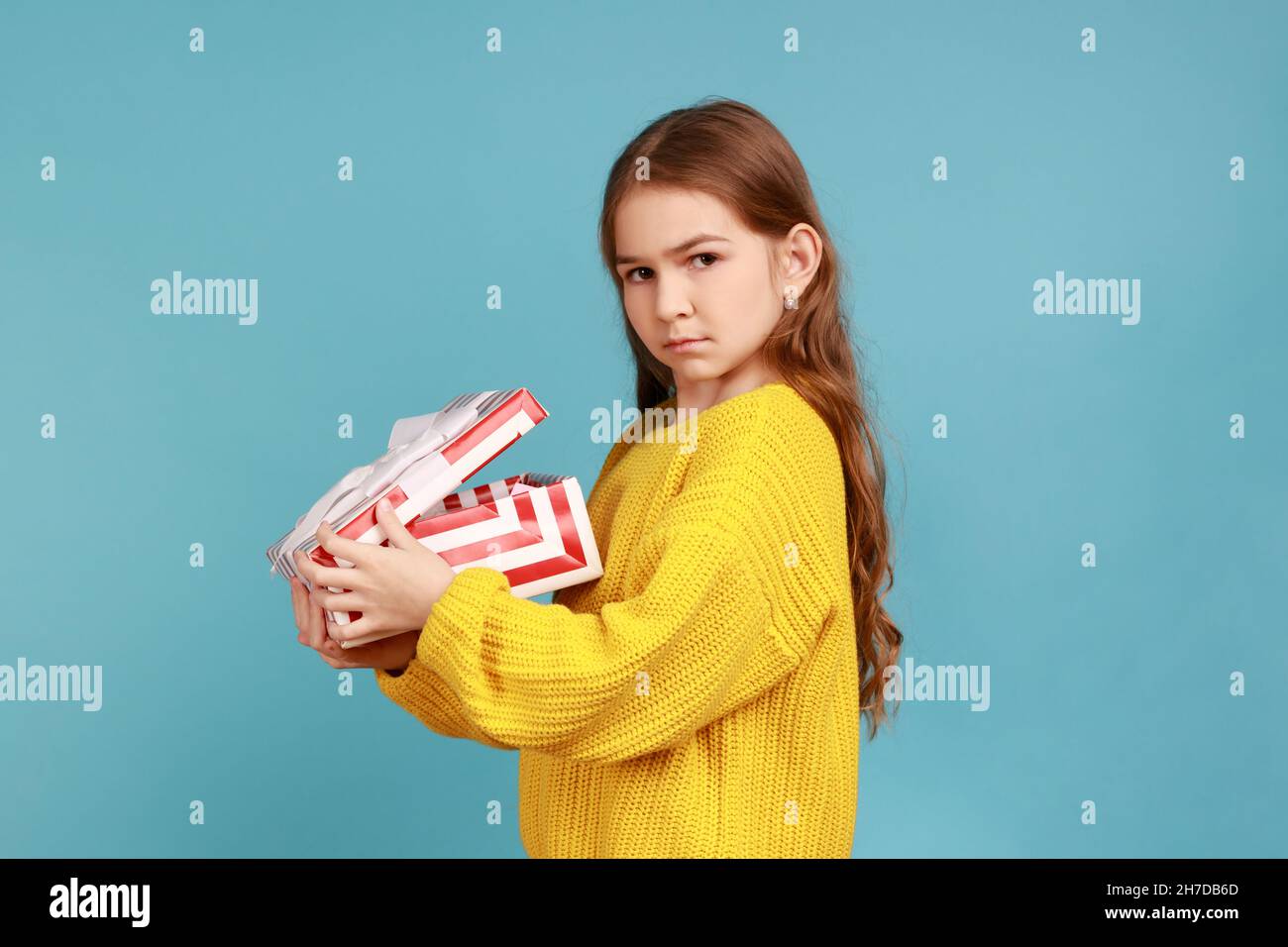 Seitenansicht des kleinen Mädchens öffnet Geschenkbox, drückt Traurigkeit aus, feiert Urlaub, enttäuscht vom Geschenk, trägt gelben lässigen Stil Pullover. Innenaufnahme des Studios isoliert auf blauem Hintergrund. Stockfoto