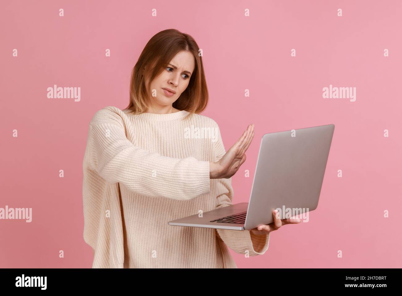 Frau zeigt Stoppgeste auf dem Laptop-Bildschirm, Warnung mit Verbotsschild beim Telefonieren in einem Videoanruf, Online-Kommunikation, trägt weißen Pullover. Innenaufnahme des Studios isoliert auf rosa Hintergrund. Stockfoto