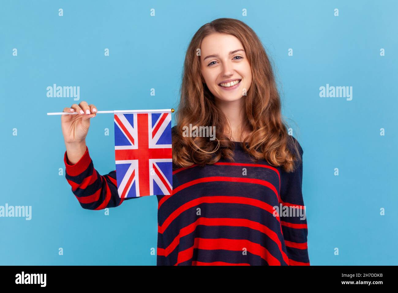 Attraktive Frau mit gestreiftem Pullover im lässigen Stil, die die Flagge einer britischen Einheit hält und den britischen Unabhängigkeitstag feiert, der isoliert auf blauem Hintergrund aufgenommen wurde. Stockfoto