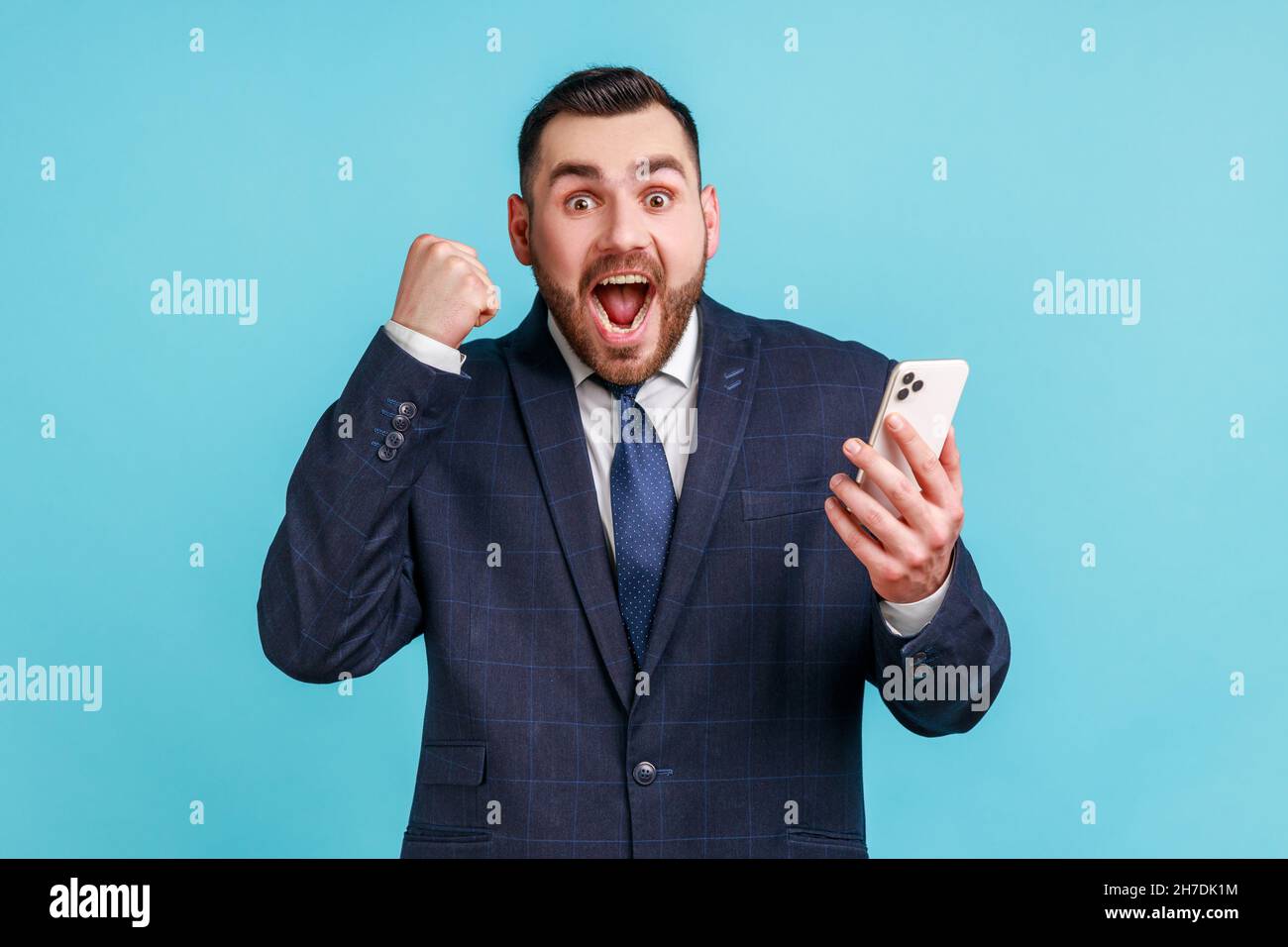 Ich bin Champion. Attraktiver Mann mit Bart trägt dunklen offiziellen Stil Anzug stehen, halten Telefon, machen Selfie und feiern den Sieg. Innenaufnahme des Studios isoliert auf blauem Hintergrund. Stockfoto