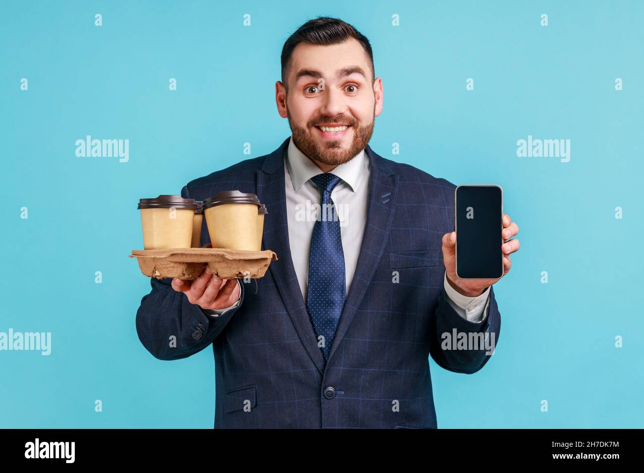 Extrem glücklich Mann trägt offiziellen Stil Anzug in den Händen halten leeren Display Smartphone und Halter mit Kaffee Pappbecher, Online-Bestellung. Innenaufnahme des Studios isoliert auf blauem Hintergrund. Stockfoto