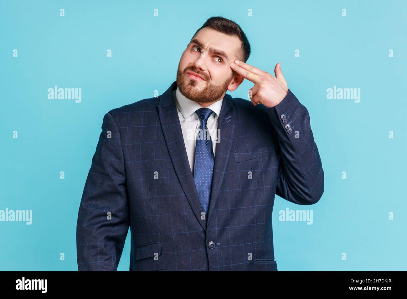 Depression, Selbstmordgestik. Porträt eines Geschäftsmannes, der einen Anzug im offiziellen Stil mit einem auf den Kopf gespitzten Fingergewehr trägt und sich mit einer Handpistole schießt. Innenaufnahme des Studios isoliert auf blauem Hintergrund. Stockfoto