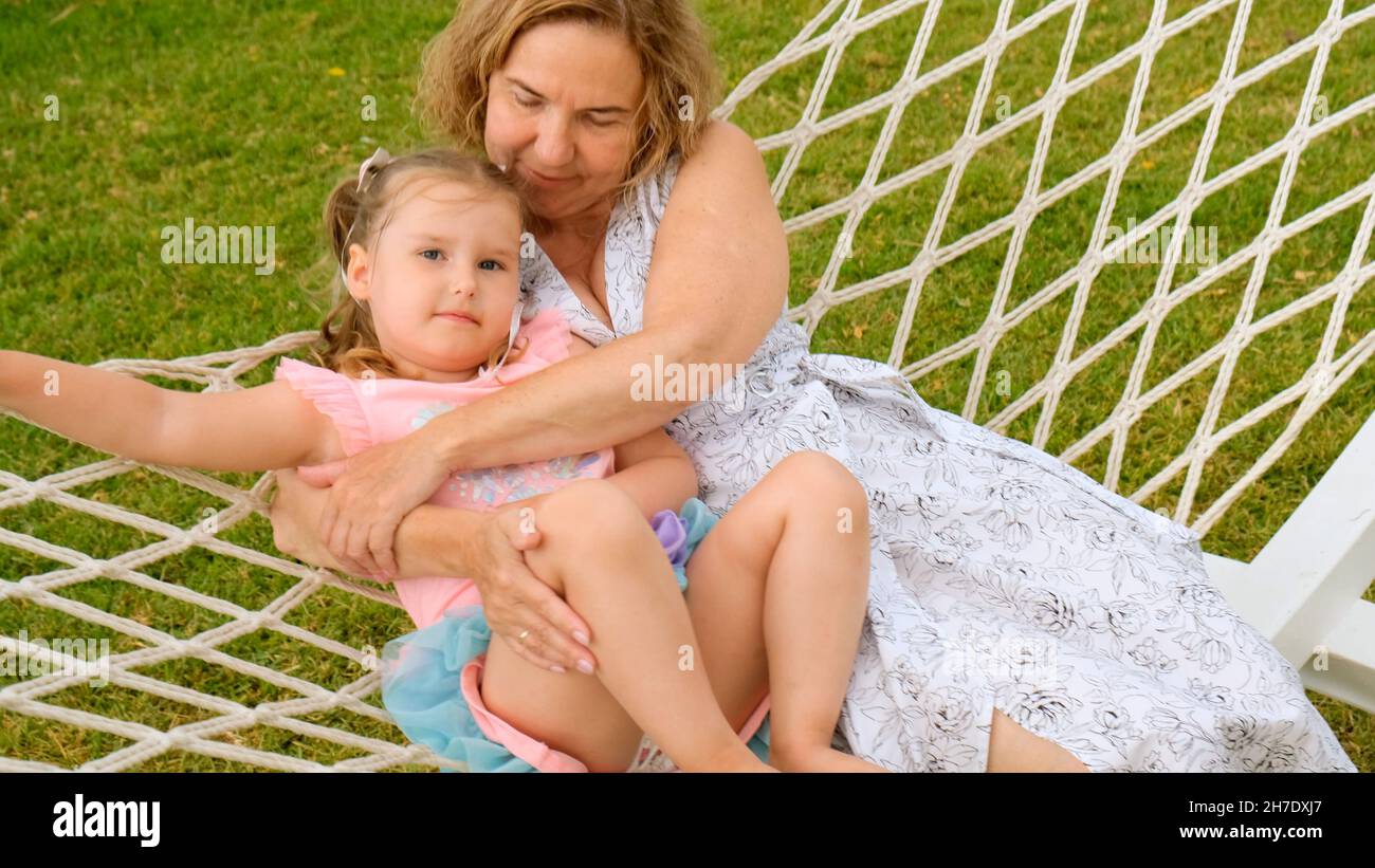 Das fröhliche Paar Großmutter und Enkelin 3 Jahre alt entspannen sich und sonnen sich in einer Hängematte auf einer grünen Wiese in ihrem Garten an einem sonnigen Sommer Stockfoto